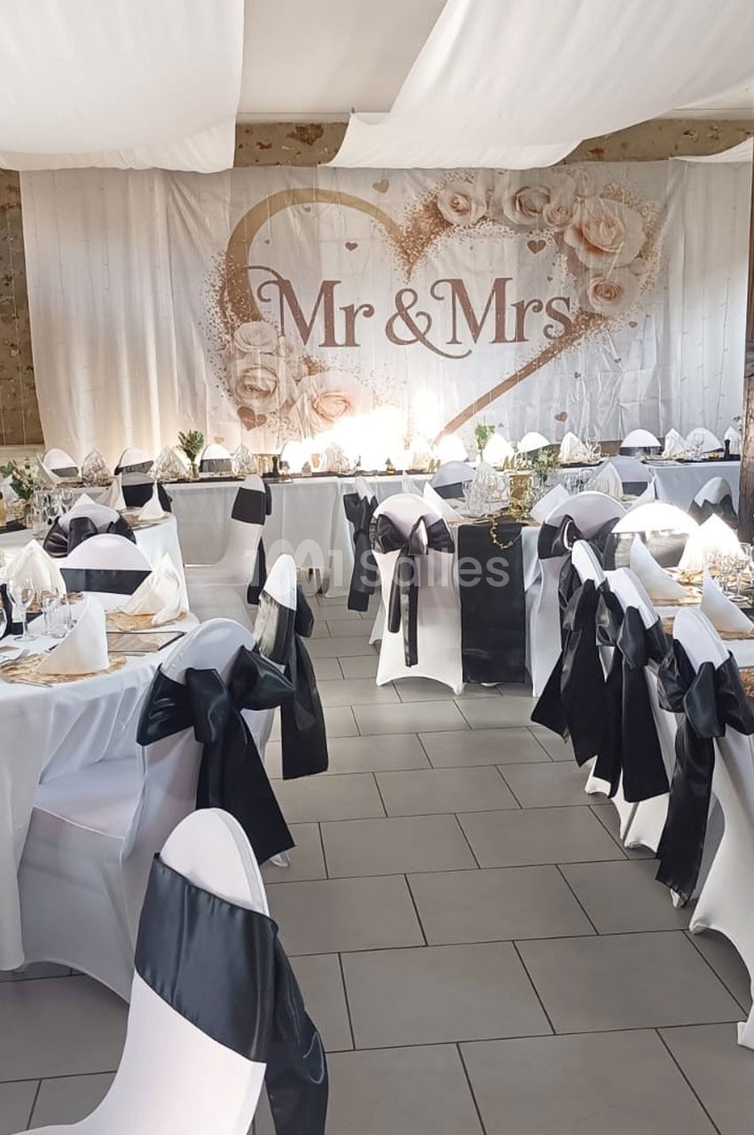 Salle de réception décorée pour un mariage, avec des tables dressées et un fond ’Mr & Mrs’ orné de fleurs.