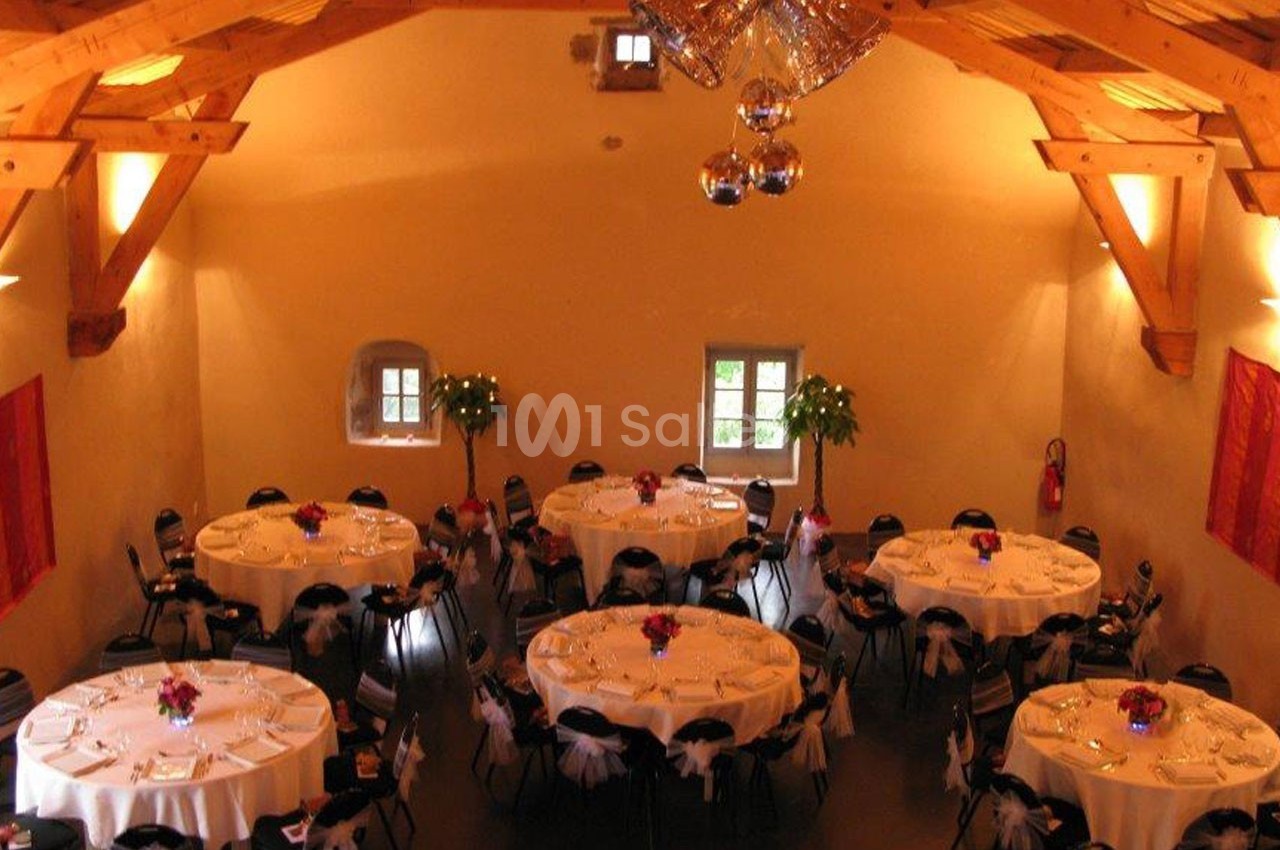 Salle de réception avec des tables rondes dressées, décorée de poutres en bois apparentes et de plantes.