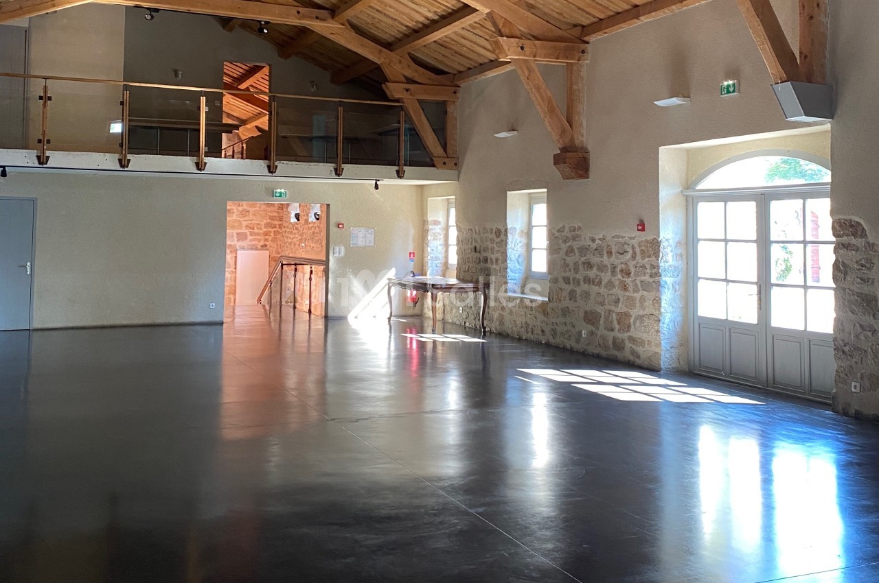 Salle lumineuse avec sol en béton poli, murs en pierre, poutres apparentes et mezzanine en bois.