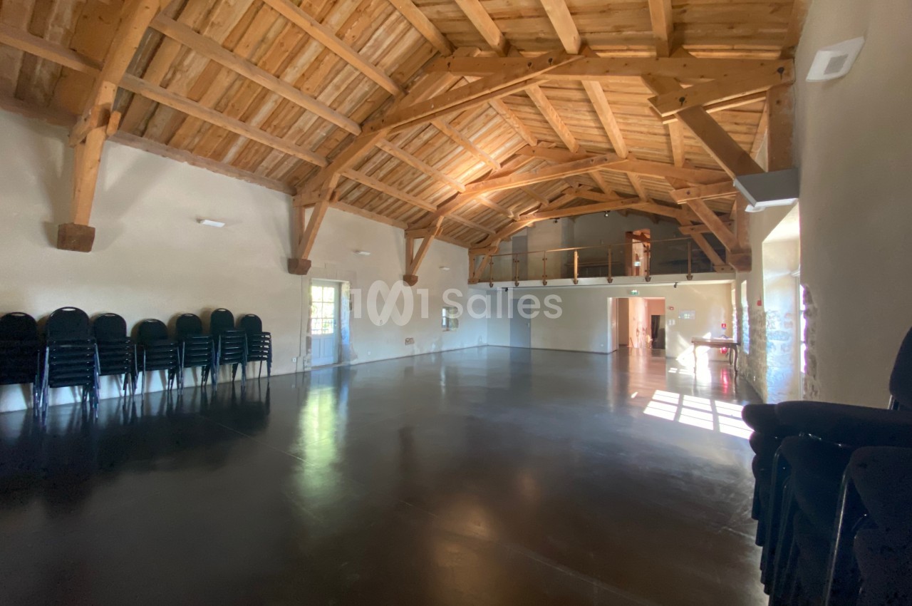Grande salle avec charpente en bois apparente, sol en béton lisse, chaises empilées sur le côté et lumière naturelle.