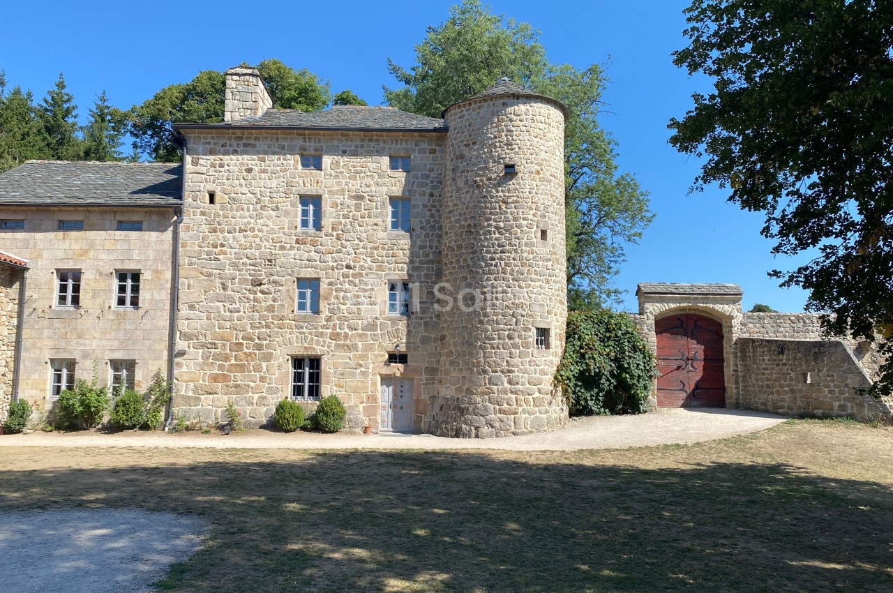 Façade en pierre d'un bâtiment historique avec une tour ronde, entouré de verdure et d'une grande porte en bois.