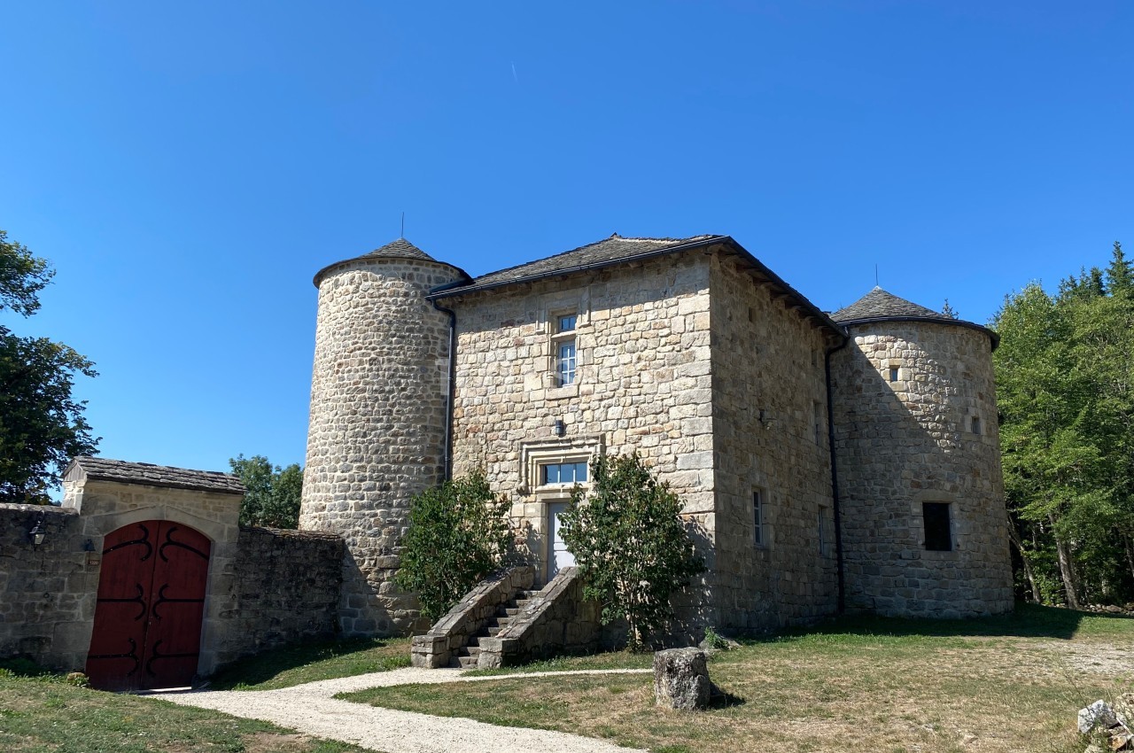 Bâtisse en pierre avec deux tours rondes, une porte rouge et un escalier, entourée de verdure sous un ciel bleu.