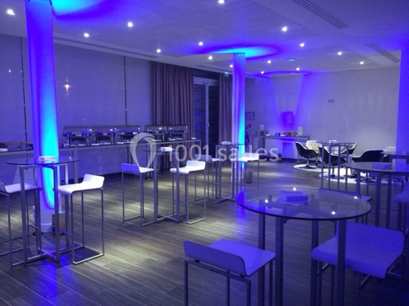 Salle moderne éclairée par des lumières bleues, avec tables hautes, tabourets blancs et buffet en arrière-plan.