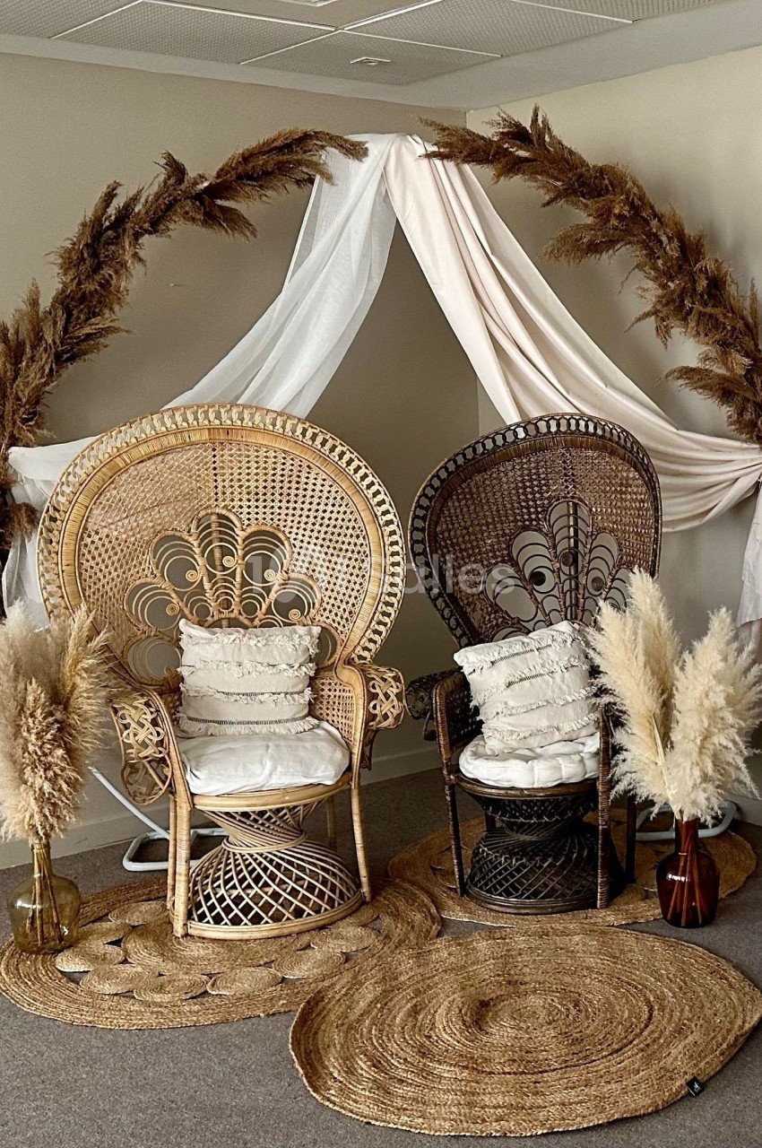 Deux fauteuils en rotin ornés de coussins, disposés sous une arche décorée de voilages et de pampas.