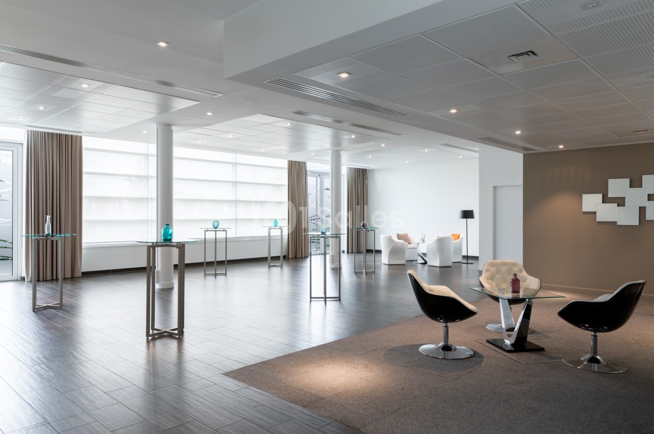 Salle lumineuse avec parquet sombre, mobilier moderne, tables hautes et fauteuils disposés dans un espace ouvert.