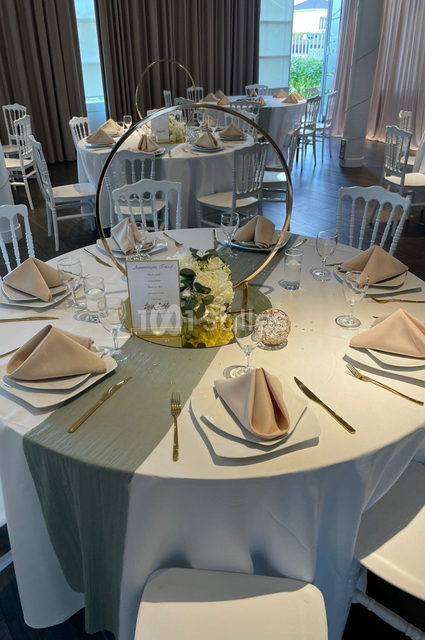Table ronde élégamment dressée avec nappes blanches, serviettes beige pliées, décoration florale et couverts dorés.