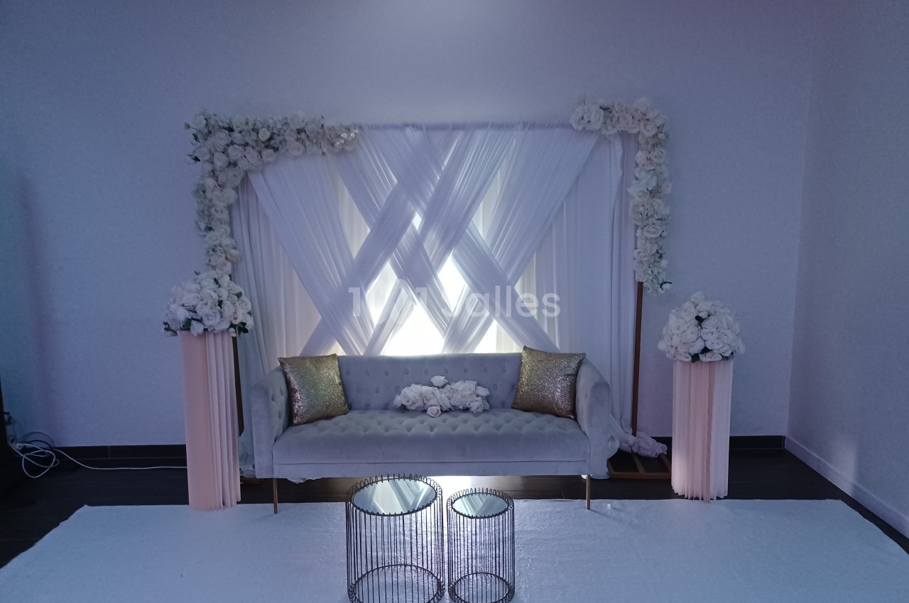 Canapé gris devant un rideau blanc décoré de fleurs blanches, avec deux tables basses et colonnes florales assorties.