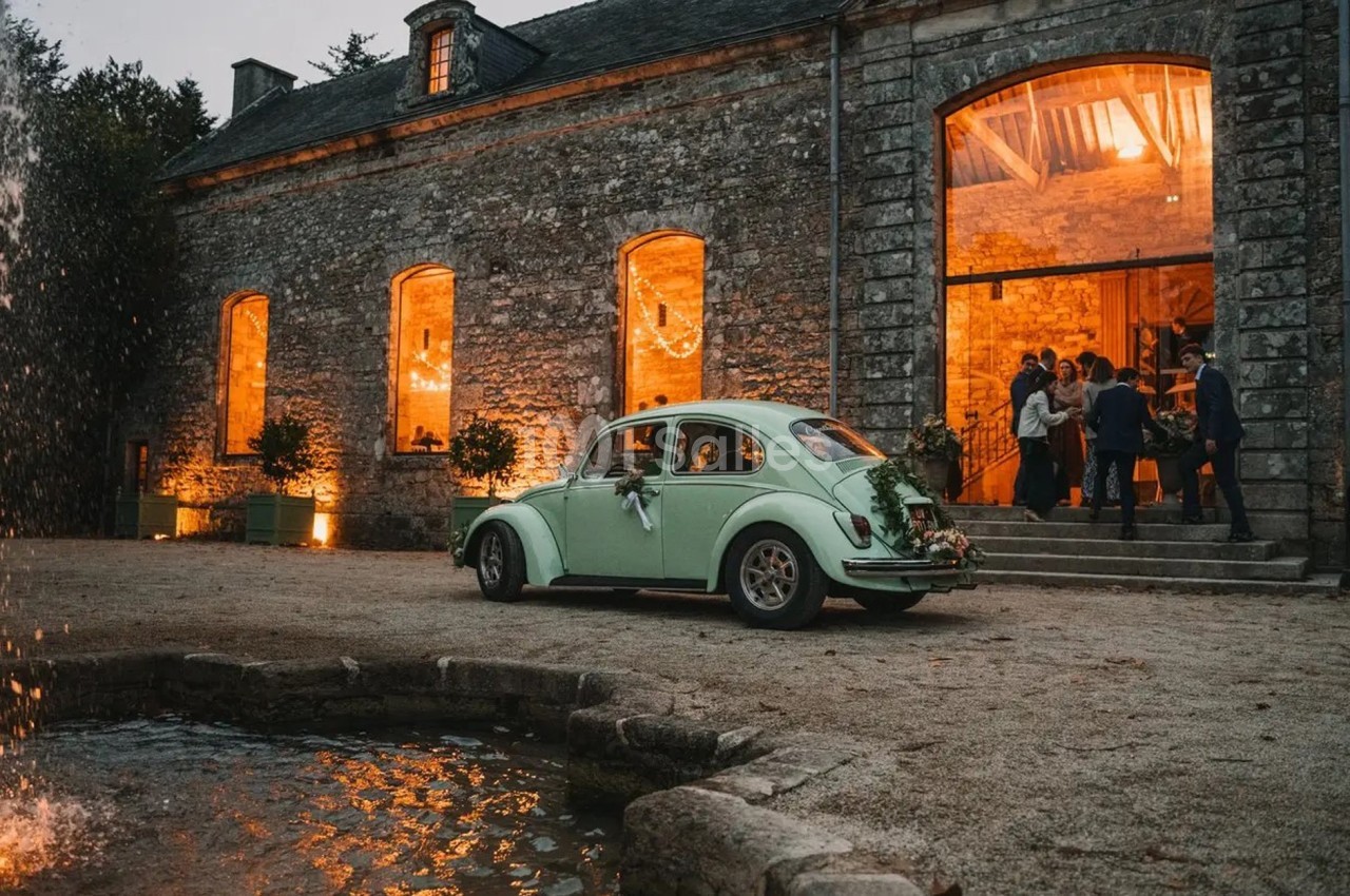 Une voiture ancienne vert clair décorée pour un mariage est garée devant un bâtiment en pierre éclairé.