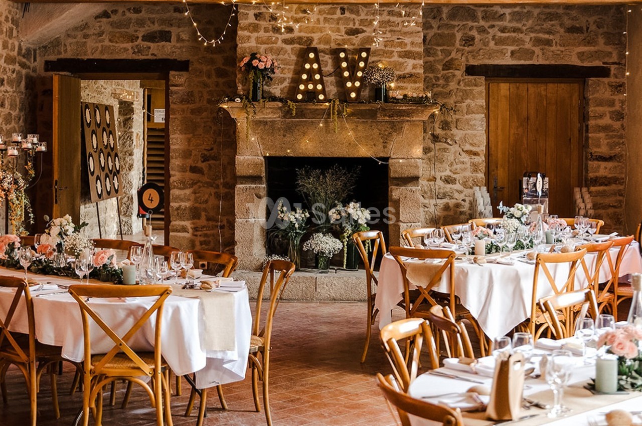 Salle de réception décorée pour un mariage, avec tables dressées, chaises en bois, fleurs et guirlandes lumineuses.