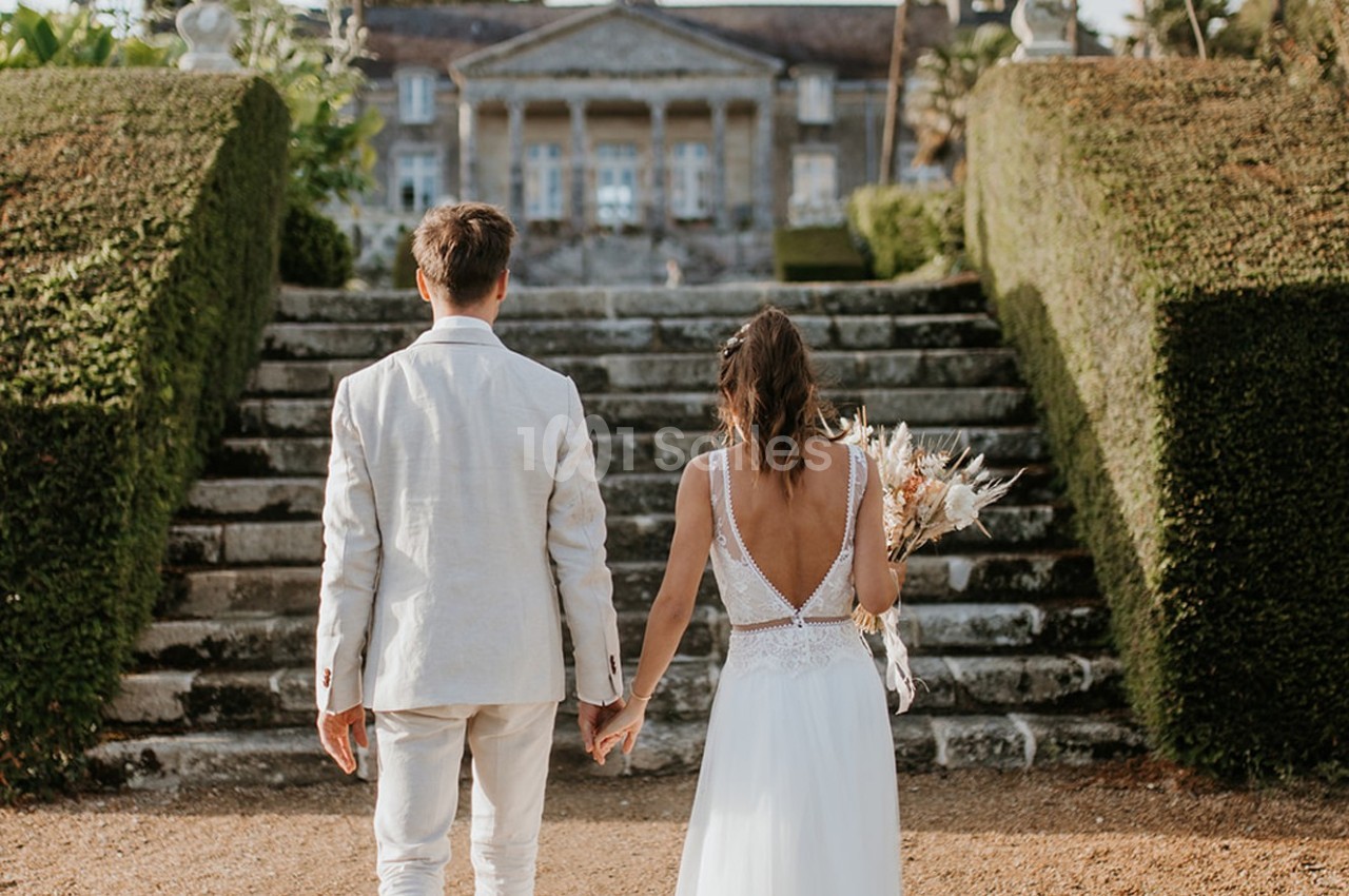 Un couple en tenue de mariage monte des marches en pierre, menant à un bâtiment historique entouré de verdure.
