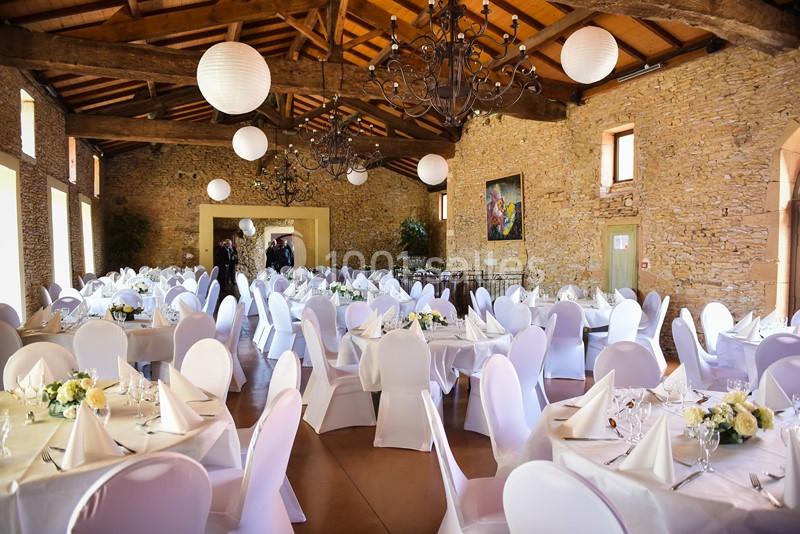 Salle de réception avec murs en pierre, tables rondes dressées, chaises blanches et lanternes suspendues au plafond en bois.