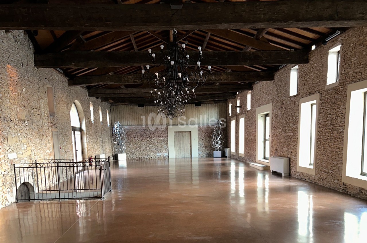 Grande salle en pierre avec plafond en poutres apparentes, lustre central et grandes fenêtres laissant entrer la lumière.