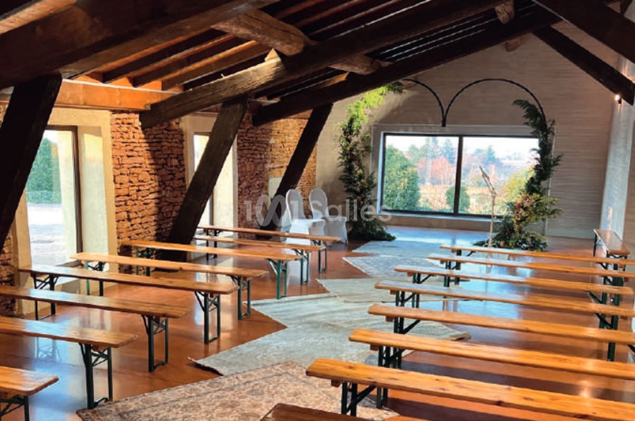 Salle lumineuse avec poutres apparentes, bancs en bois alignés et grande baie vitrée donnant sur un paysage arboré.