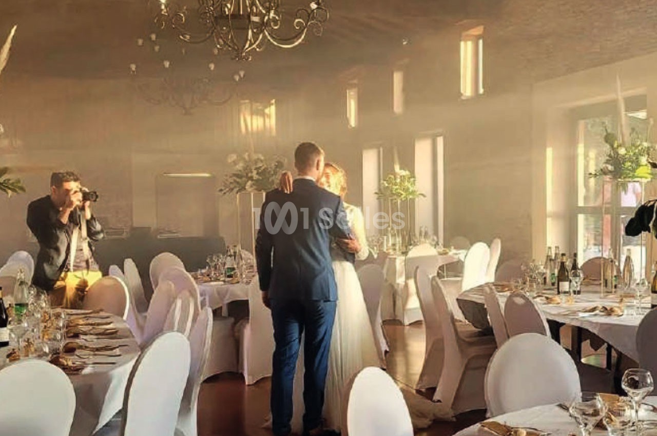 Un couple danse dans une salle de réception lumineuse, entourée de tables décorées et d'un photographe capturant la scène.