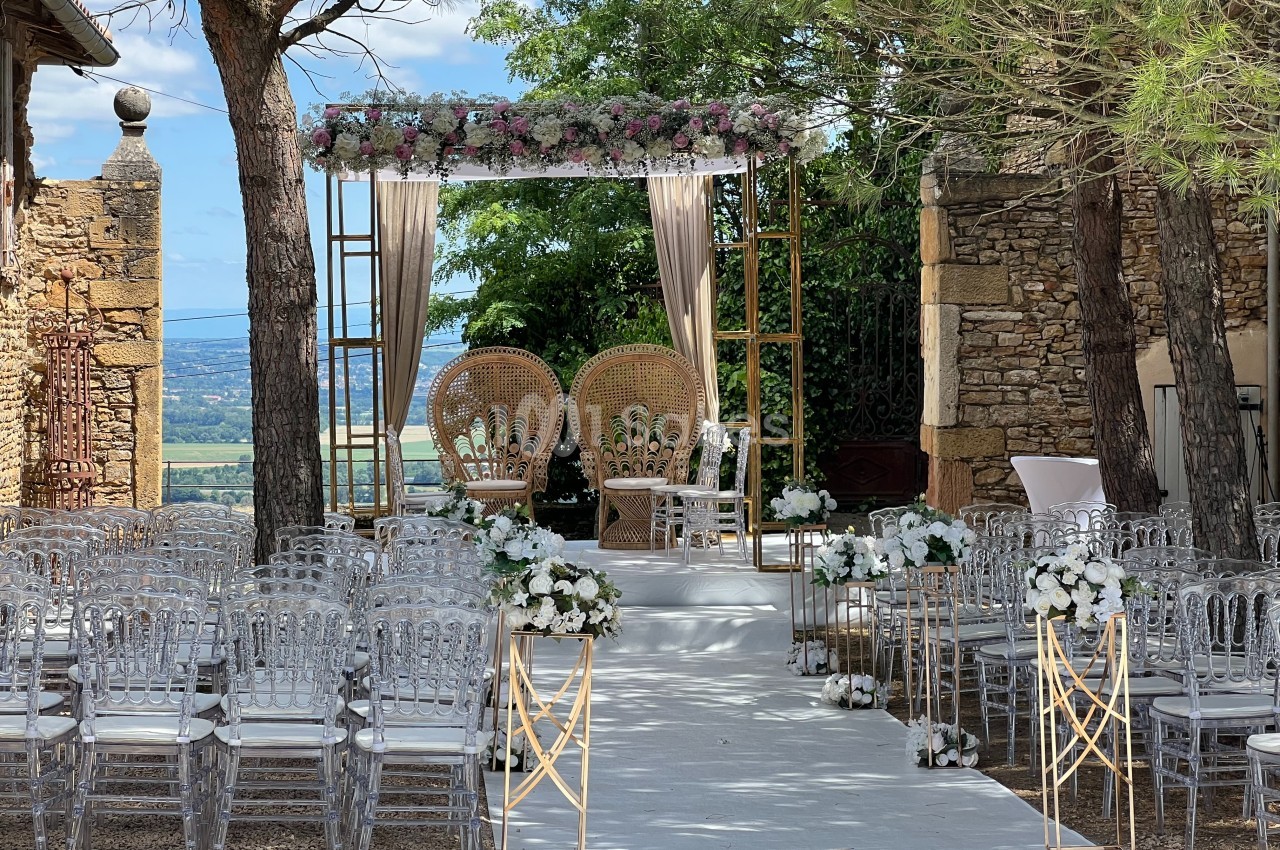 Allée décorée de fleurs menant à une arche ornée de voilages et de fleurs, entourée de chaises transparentes.