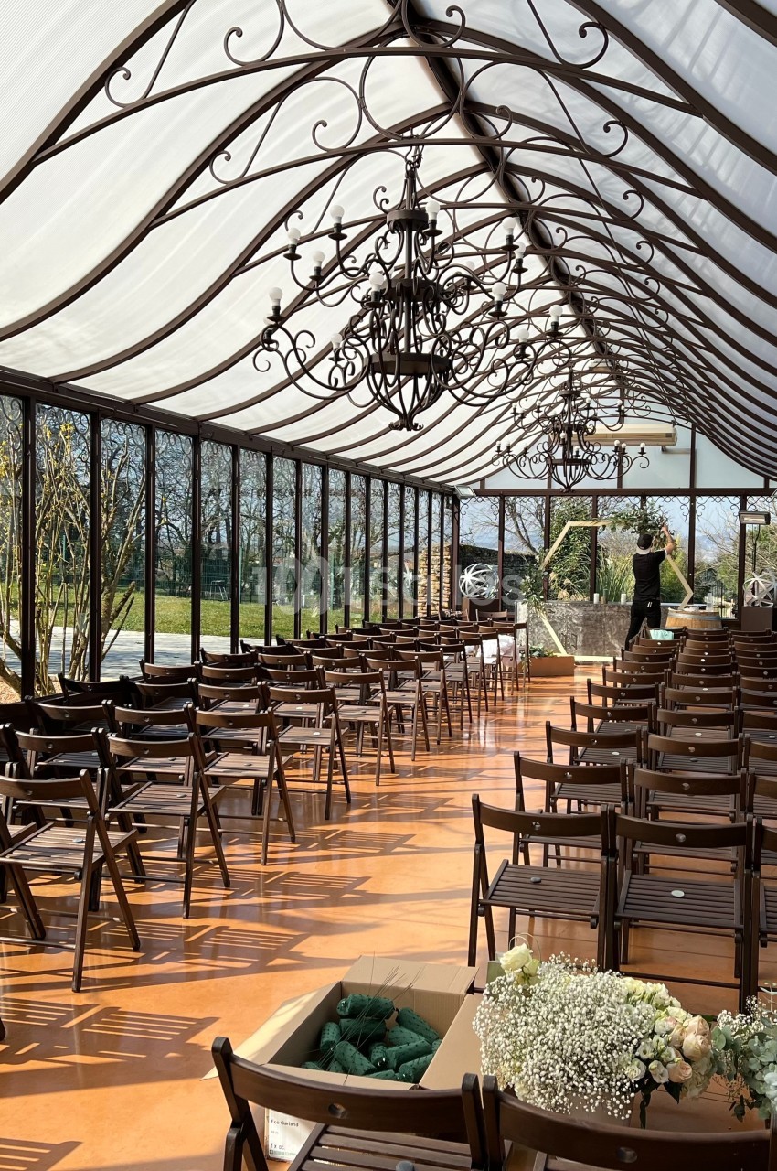 Salle lumineuse avec chaises alignées sous une verrière, décorée de lustres en fer forgé et fleurs.