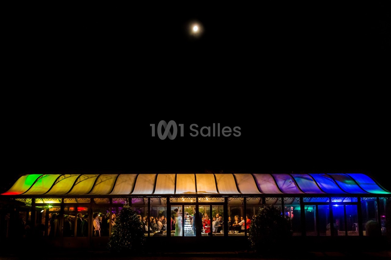 Vue nocturne d'une salle vitrée éclairée par des lumières colorées, avec des personnes visibles à l'intérieur.