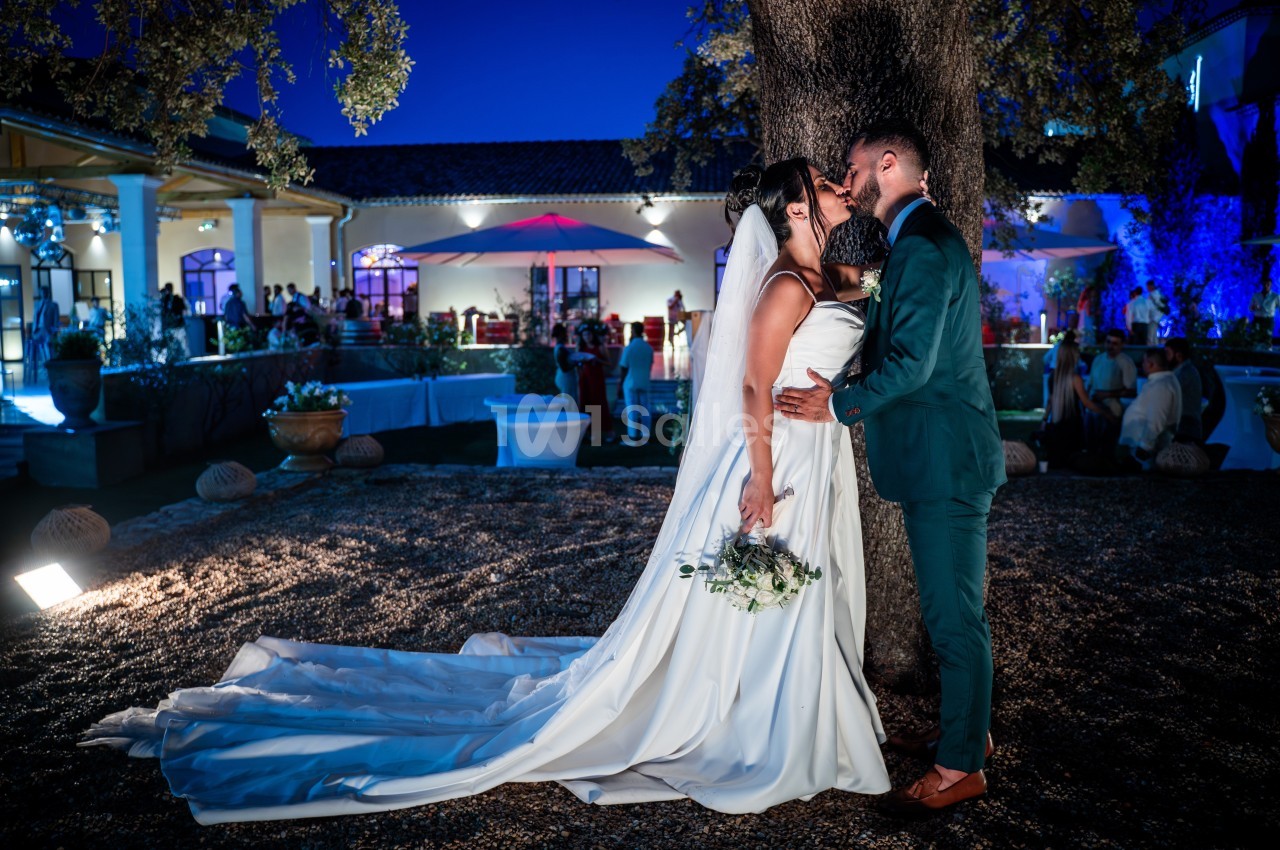 Un couple de mariés s'embrasse sous un arbre, dans un jardin éclairé en soirée près d'une salle de réception.