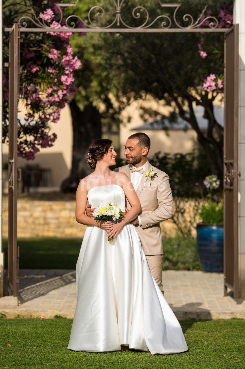 Un couple en tenue de mariage pose dans un jardin fleuri, encadré par une arche en fer forgé.