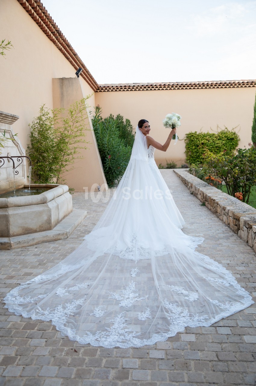 Une mariée en robe blanche avec une longue traîne tient un bouquet dans une cour pavée bordée de verdure.