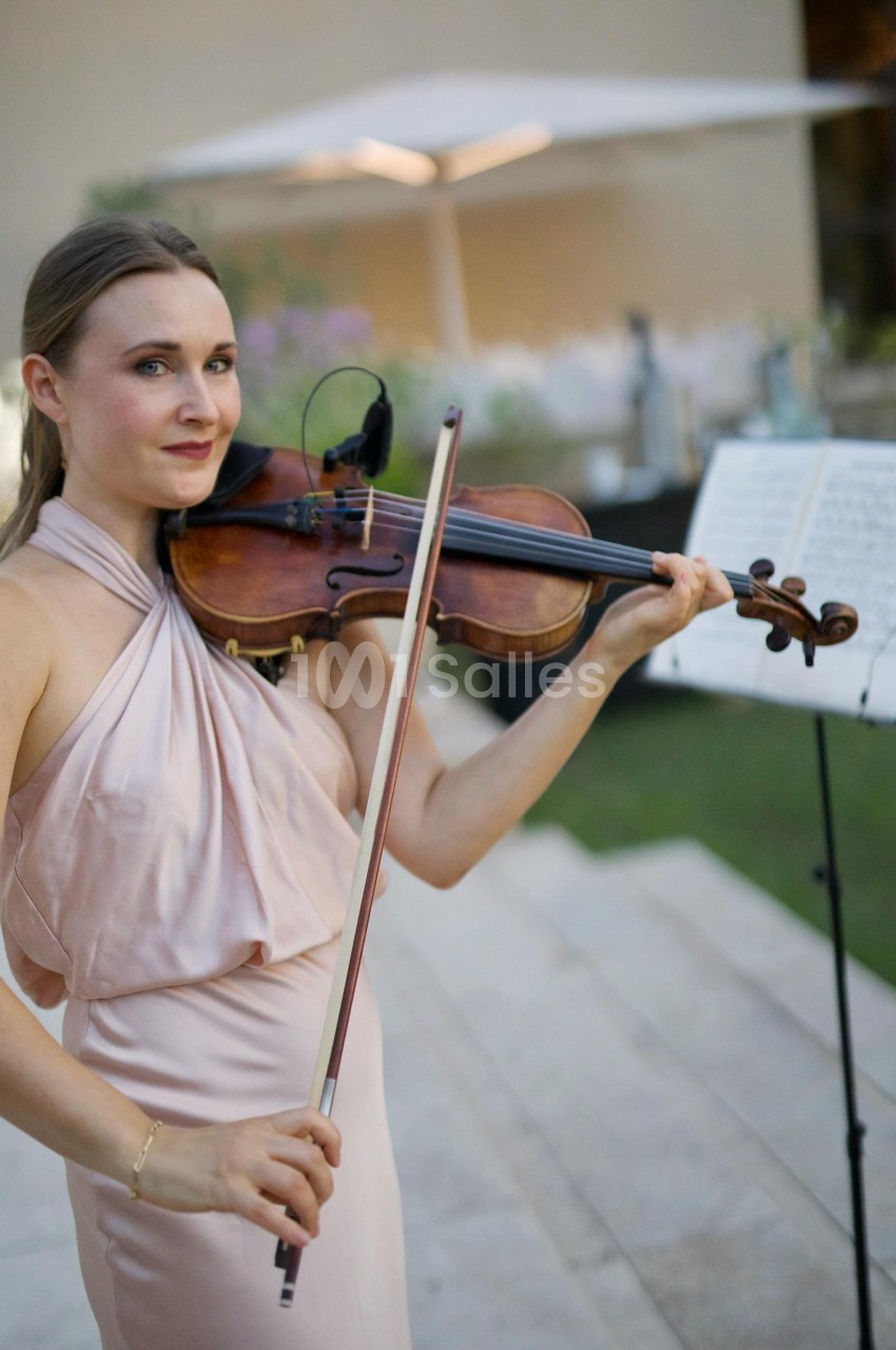 Une musicienne en robe rose joue du violon en extérieur, avec une partition sur un pupitre en arrière-plan.