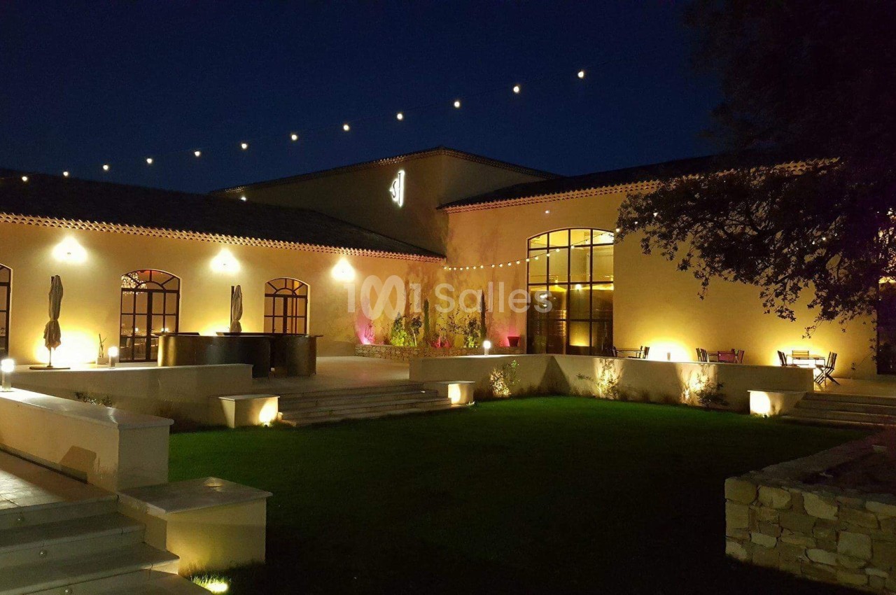 Bâtiment éclairé de nuit avec guirlandes lumineuses, grandes fenêtres et jardin aménagé au premier plan.