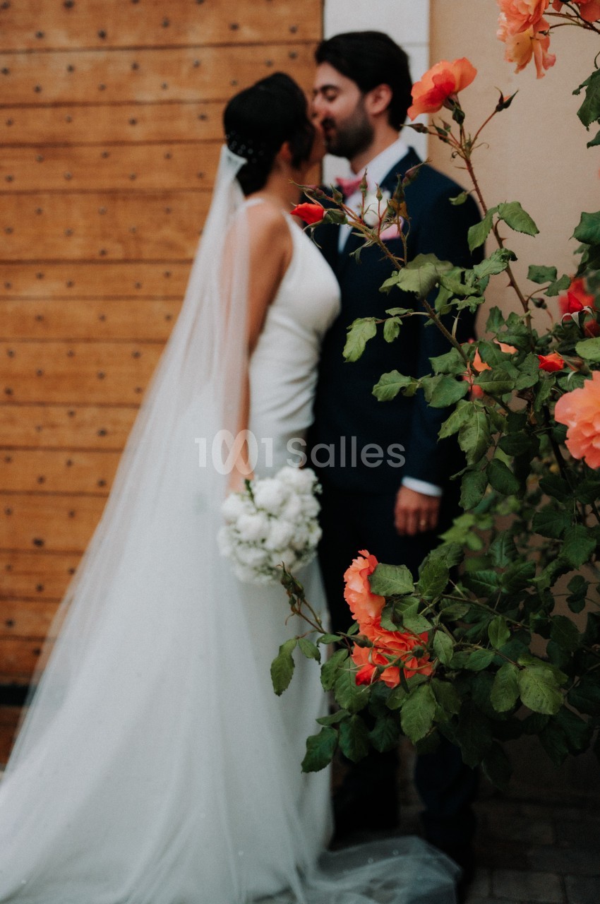 Un couple de mariés s'embrasse devant une porte en bois, avec des fleurs orangées au premier plan.