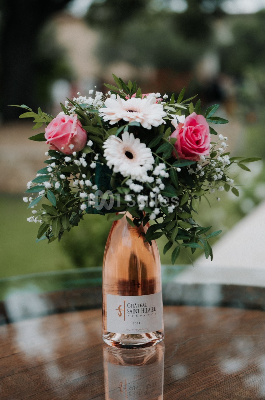 Bouquet de fleurs roses et blanches dans une bouteille de vin rosé posée sur une table en bois, en extérieur.