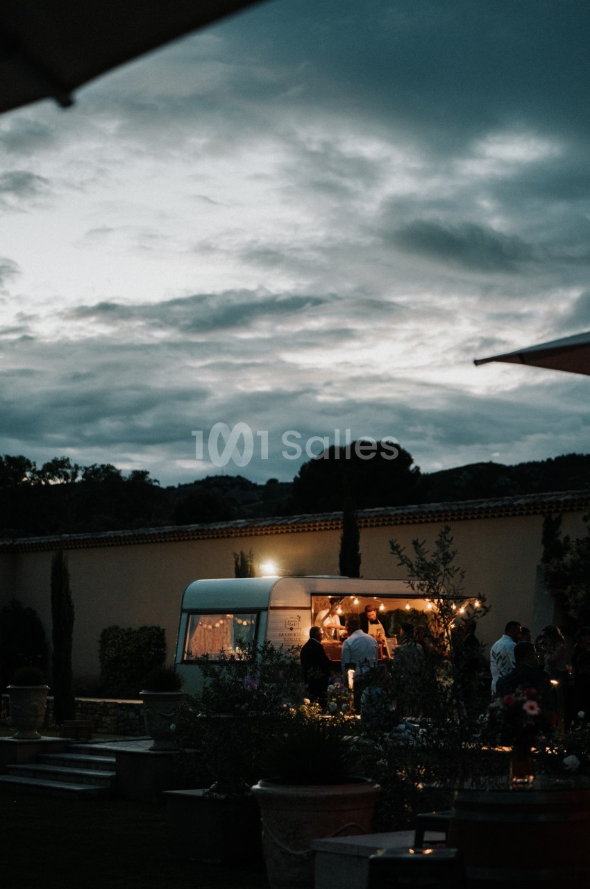 Un food truck éclairé dans un jardin au crépuscule, entouré de plantes et de quelques personnes.