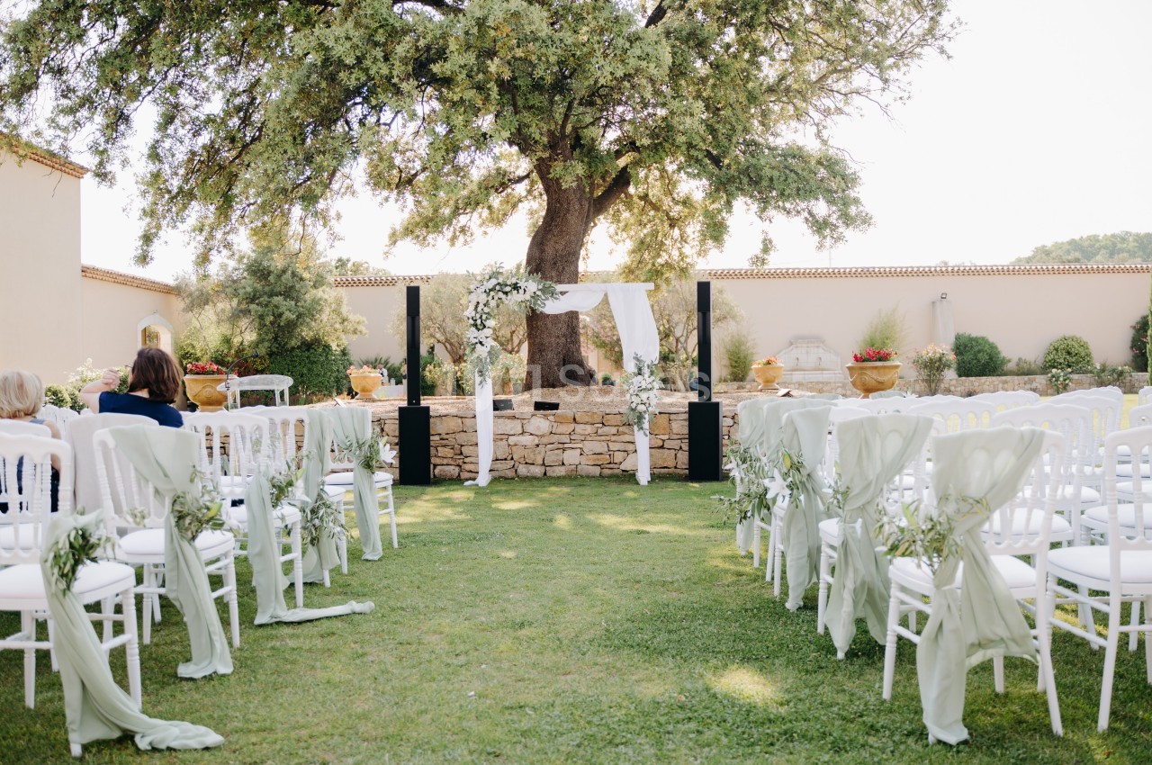 Chaises blanches décorées alignées face à une arche de mariage sous un grand arbre dans un jardin ensoleillé.