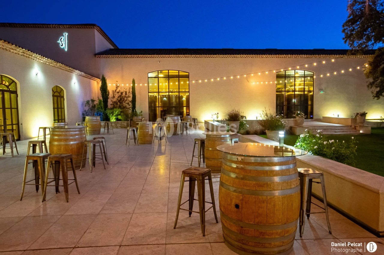 Terrasse éclairée en soirée avec tables en tonneaux, chaises métalliques et guirlandes lumineuses suspendues.