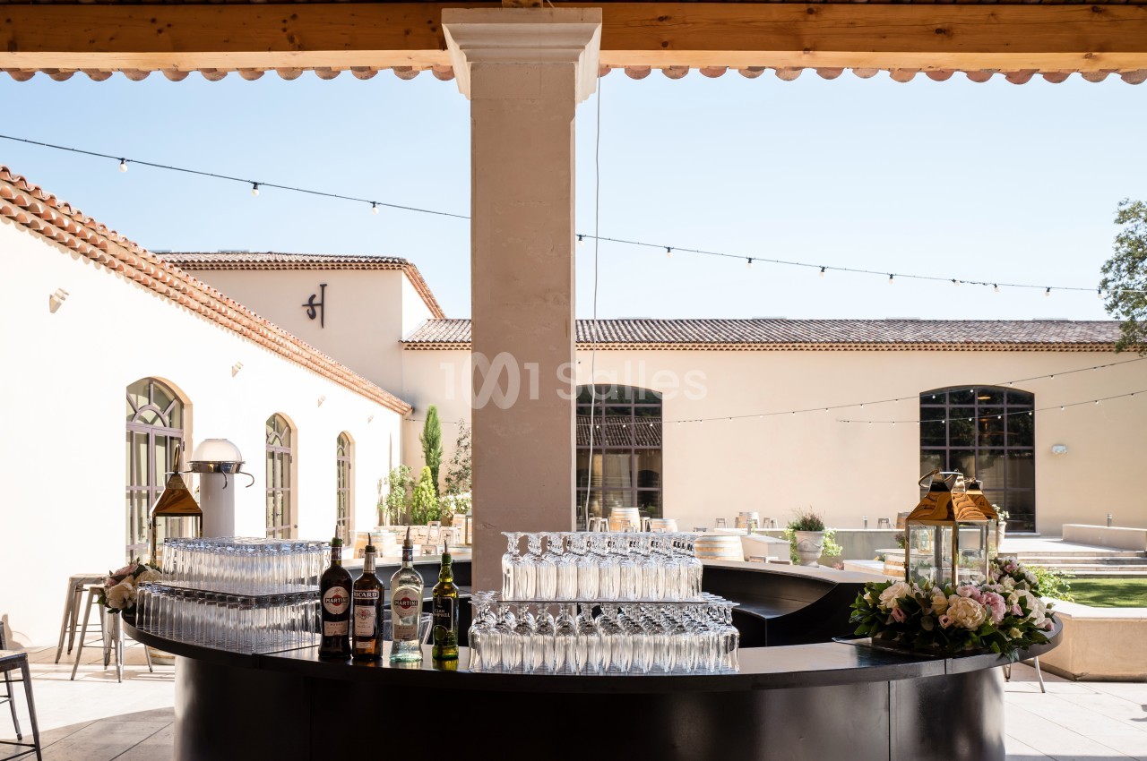 Bar extérieur avec verres empilés, bouteilles et décor floral, situé dans une cour ensoleillée avec bâtiments en arrière…