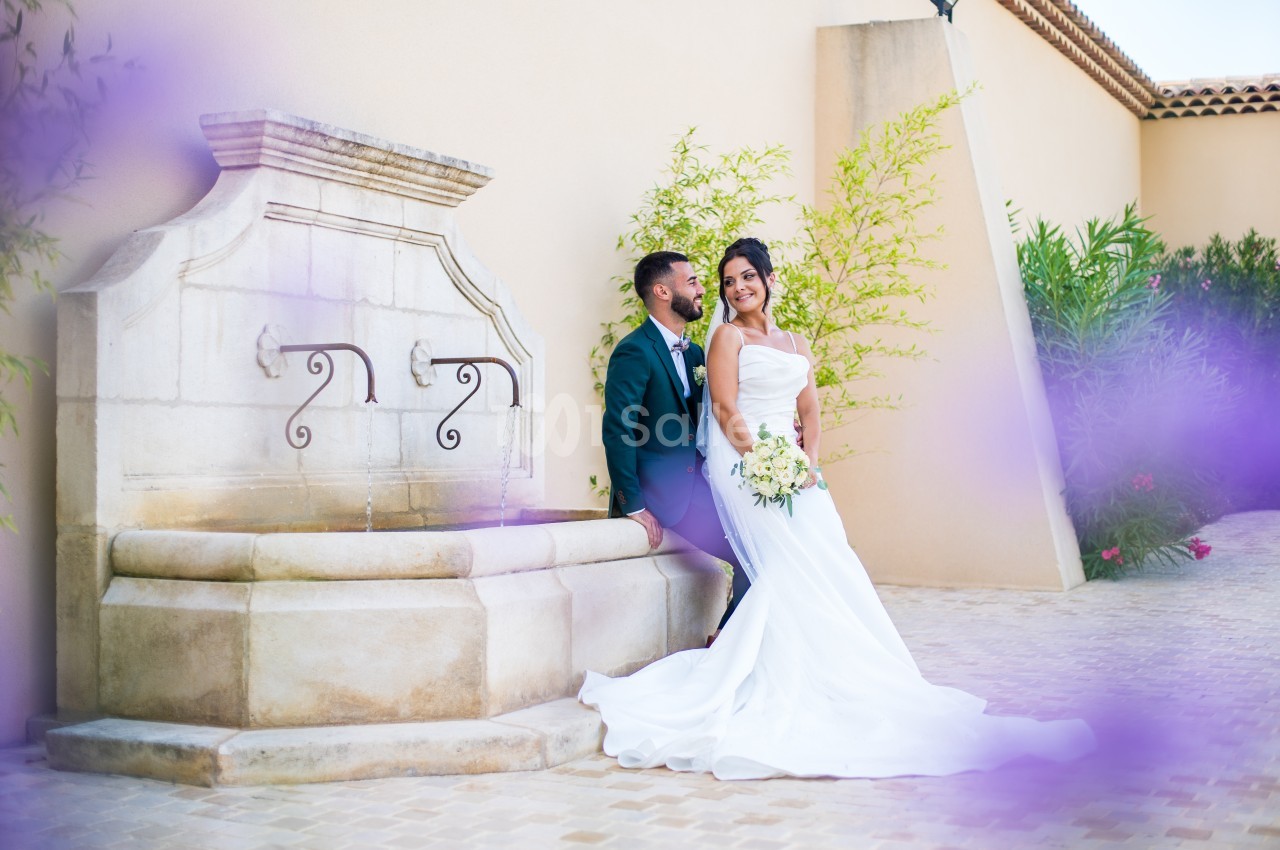 Un couple de mariés pose près d'une fontaine en pierre, entouré de végétation et de fleurs violettes floues au premier plan.