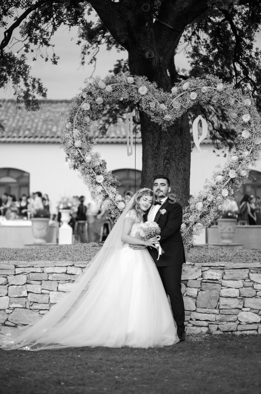 Un couple de mariés pose sous un arbre décoré d'un grand cœur floral, avec des invités en arrière-plan flou.