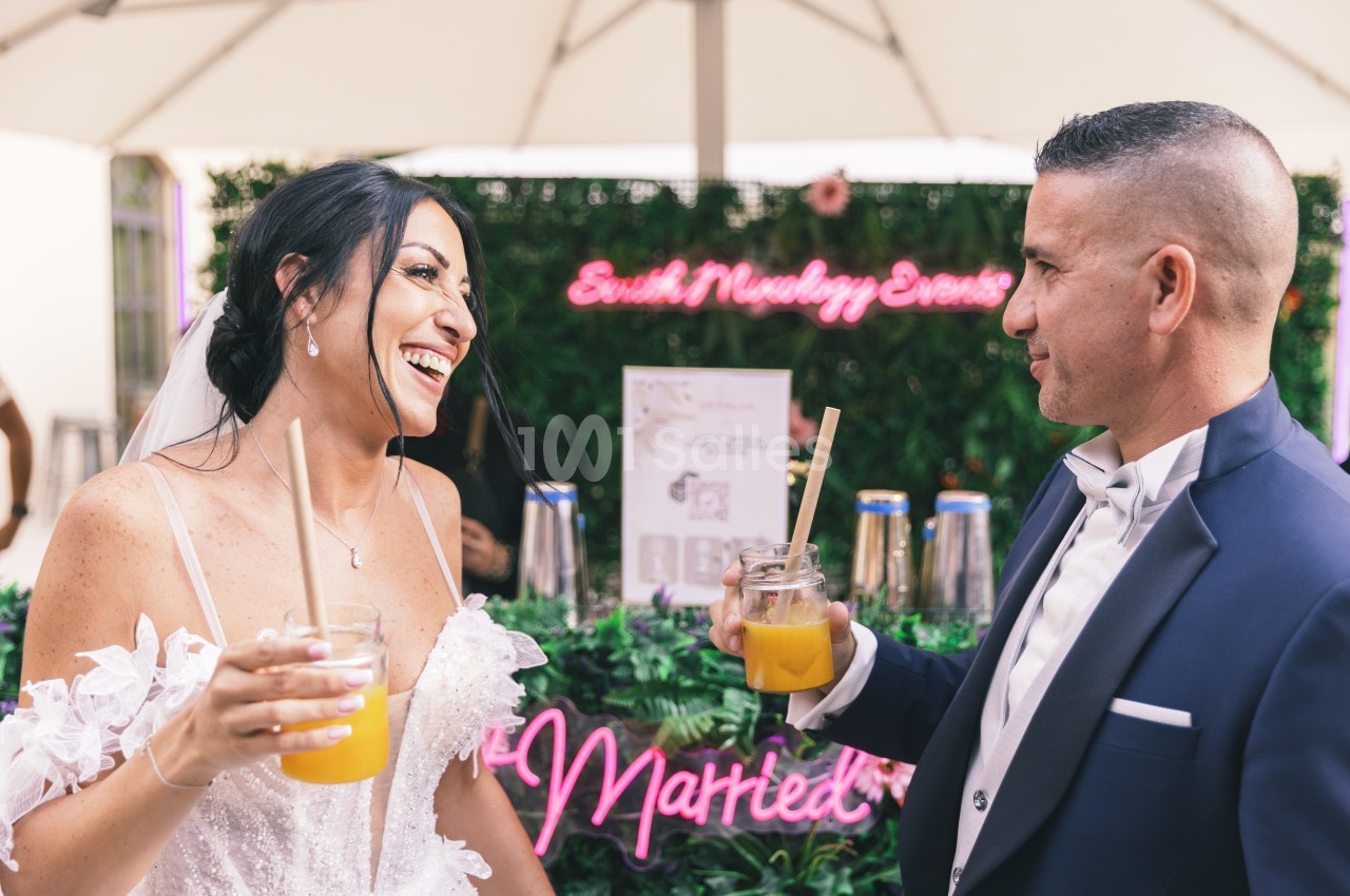 Un couple en tenue de mariage sourit en trinquant avec des boissons devant un décor floral et lumineux.