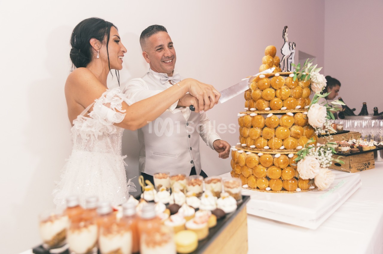 Un couple de mariés coupe une pièce montée de choux décorée de fleurs, entourée de desserts sur une table.