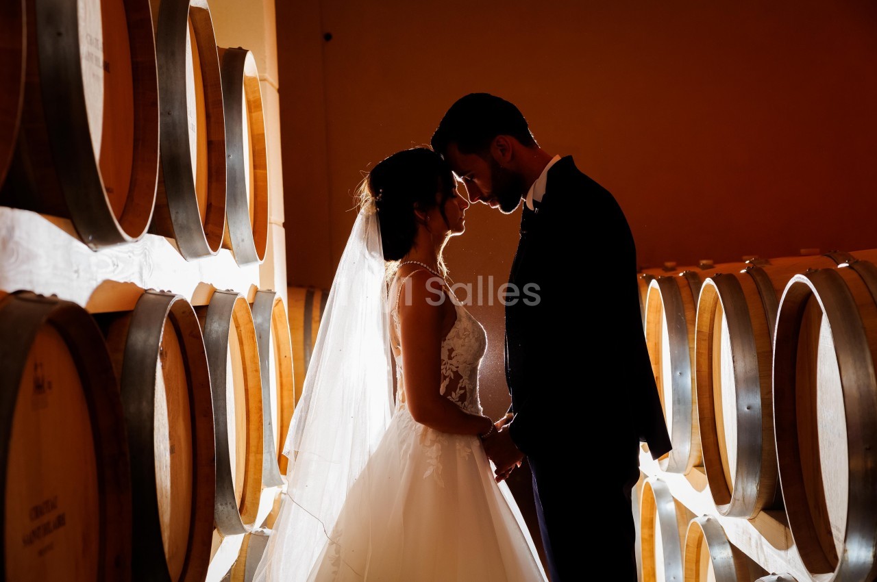 Un couple en tenue de mariage se tient face à face dans une cave entourée de tonneaux en bois.