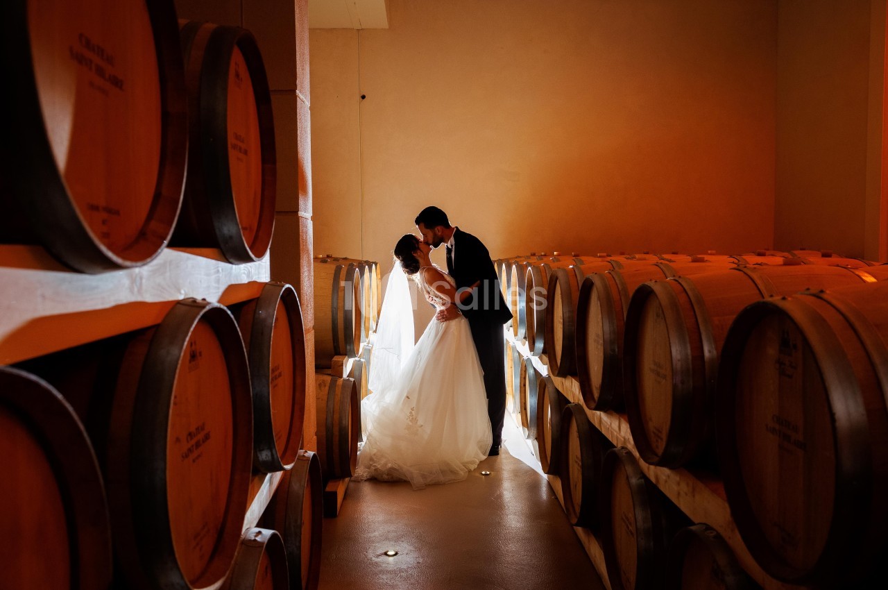Un couple en tenue de mariage s'embrasse dans une cave à vin éclairée par une lumière douce.