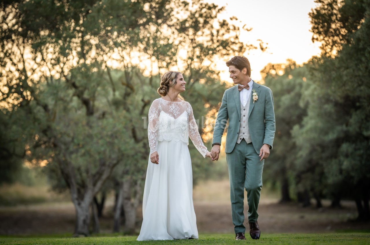 Un couple en tenue de mariage marche main dans la main sur une pelouse, entouré d'arbres au coucher du soleil.