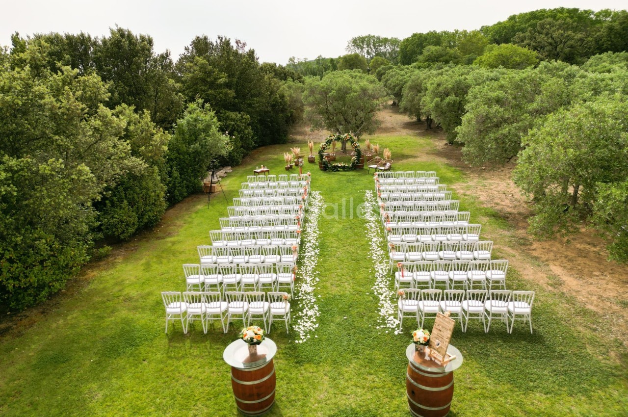 Chaises blanches alignées en extérieur pour une cérémonie, avec une arche fleurie au fond et des arbres environnants.