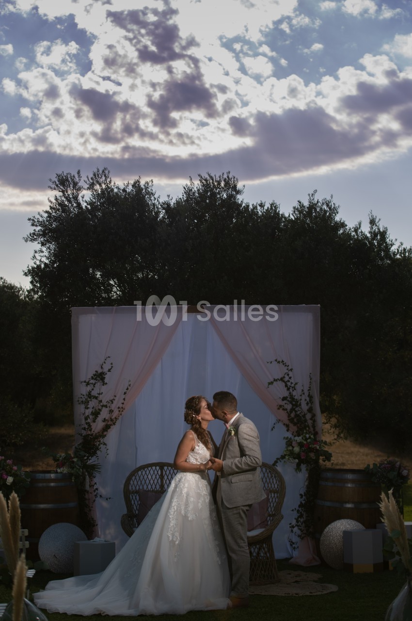 Un couple de mariés s'embrasse devant une arche décorée de voilages et de fleurs, en extérieur sous un ciel nuageux.