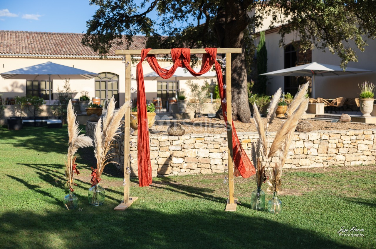 Arche en bois décorée de tissu rouge et de pampas, installée sur une pelouse devant un bâtiment en pierre.