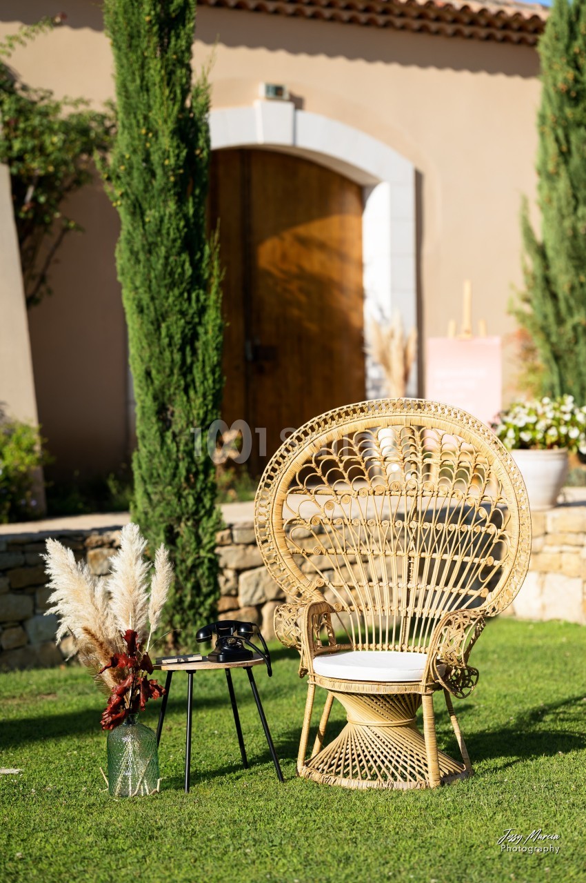 Fauteuil en rotin avec table basse et vase de pampas, installé sur une pelouse devant une maison en pierre.