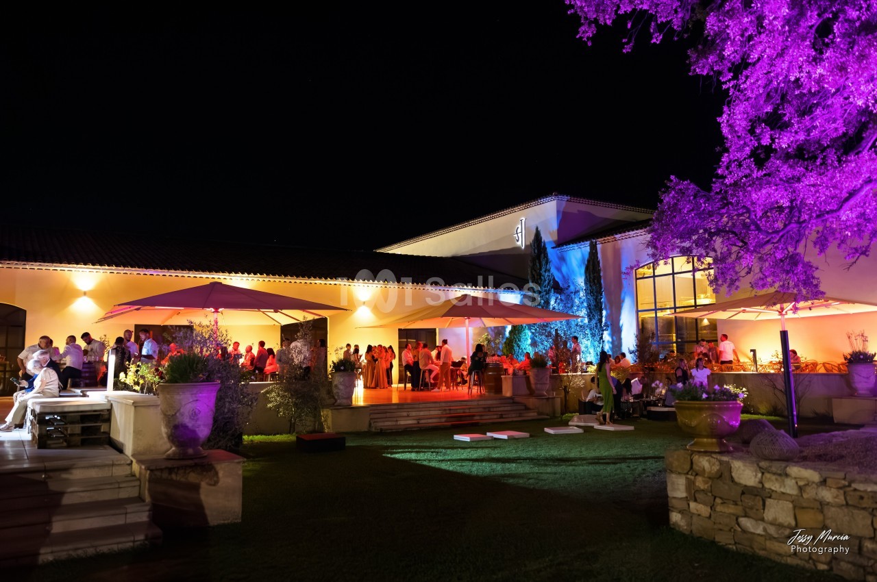 Vue nocturne d'une réception en extérieur avec invités, éclairages colorés et parasols près d'un bâtiment moderne.