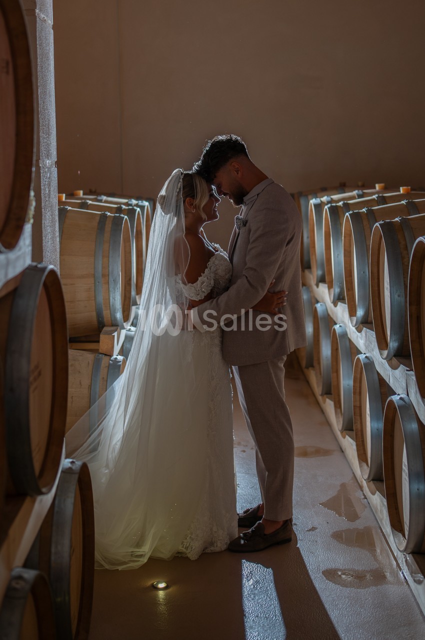 Un couple de mariés s'enlace tendrement dans une cave à vin éclairée par une lumière douce.