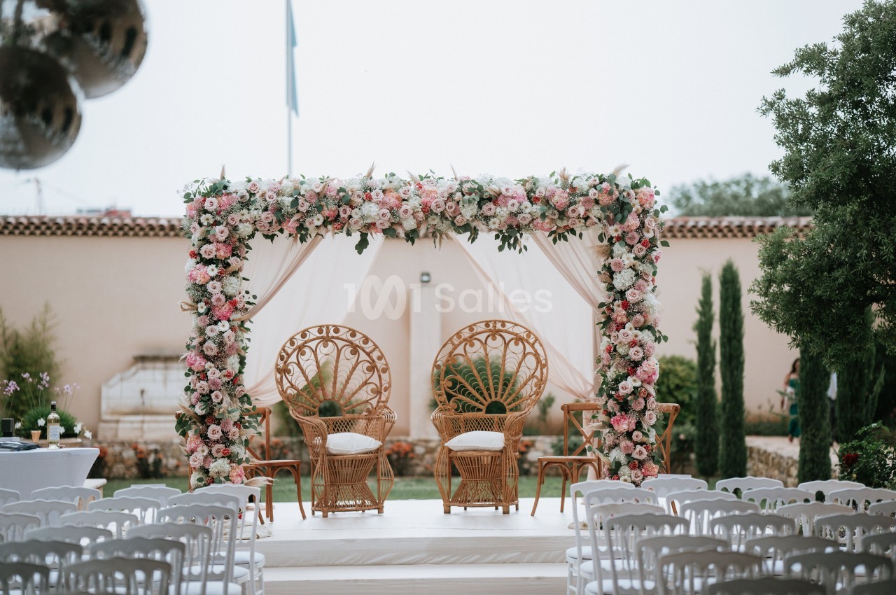 Arche florale ornée de roses et chaises en rotin disposées pour une cérémonie en extérieur.