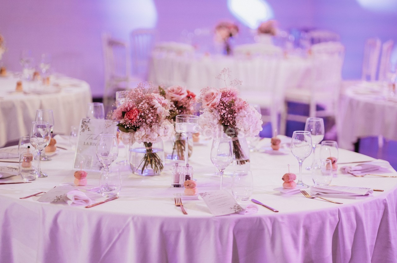 Tables décorées avec des bouquets de fleurs roses et blanches, entourées de chaises, dans une ambiance lumineuse douce.