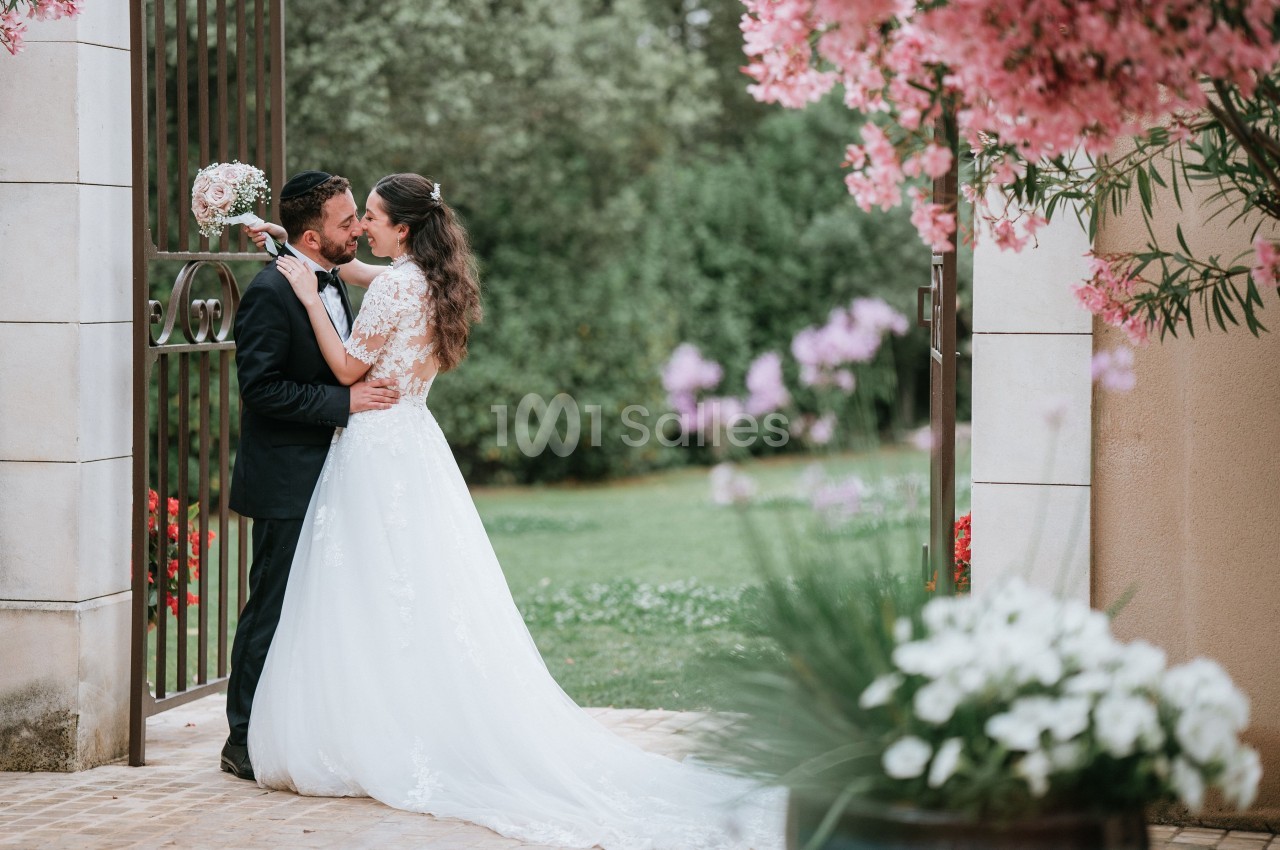 Un couple de mariés s'embrasse devant une porte en fer forgé, entouré de fleurs et d'un jardin verdoyant.