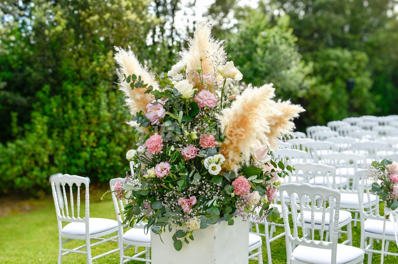 Arrangement floral avec roses, pampas et feuillage, placé devant des chaises blanches alignées dans un jardin.