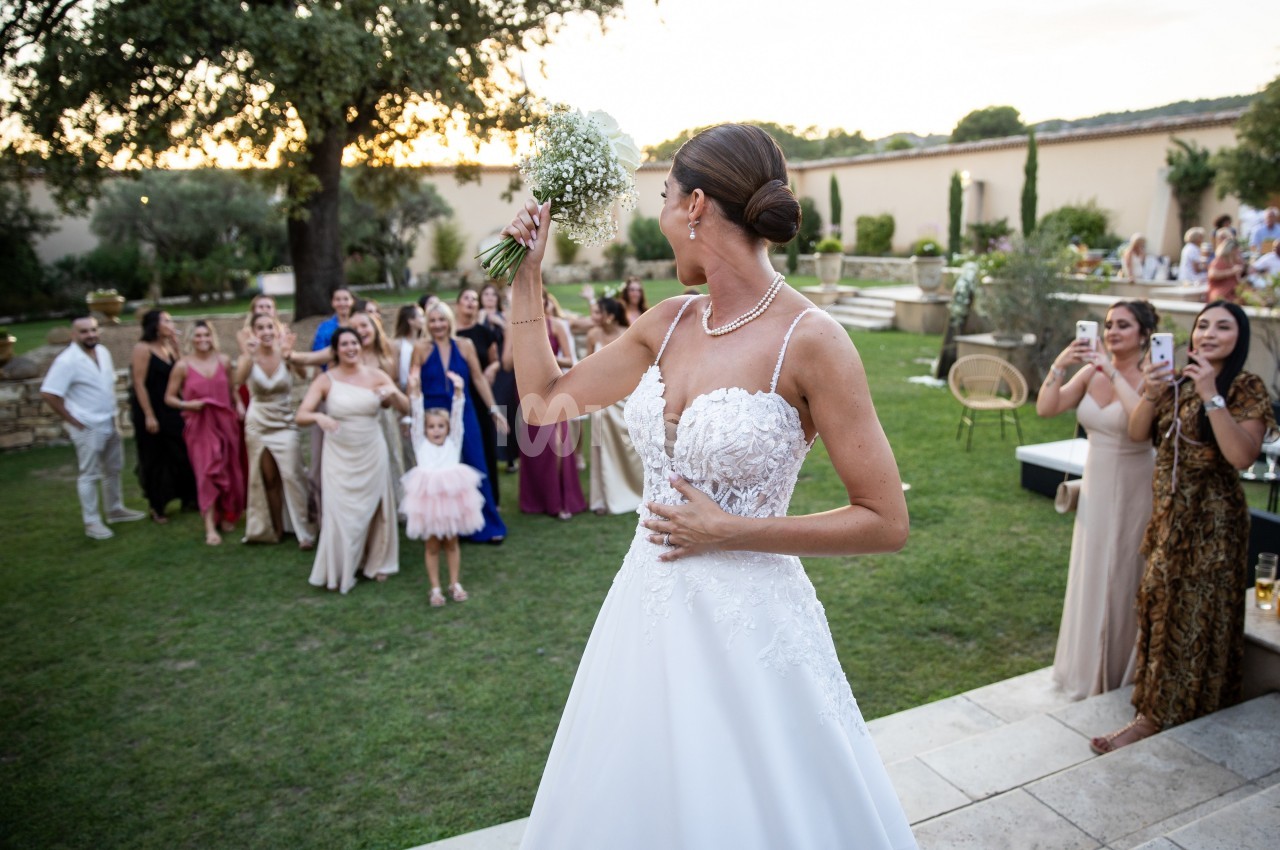 Une mariée en robe blanche lance son bouquet à un groupe de femmes rassemblées dans un jardin.